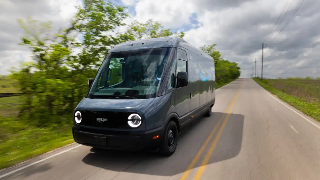 A modern electric cargo van is driving on a road with lush greenery on both sides under a cloudy sky.