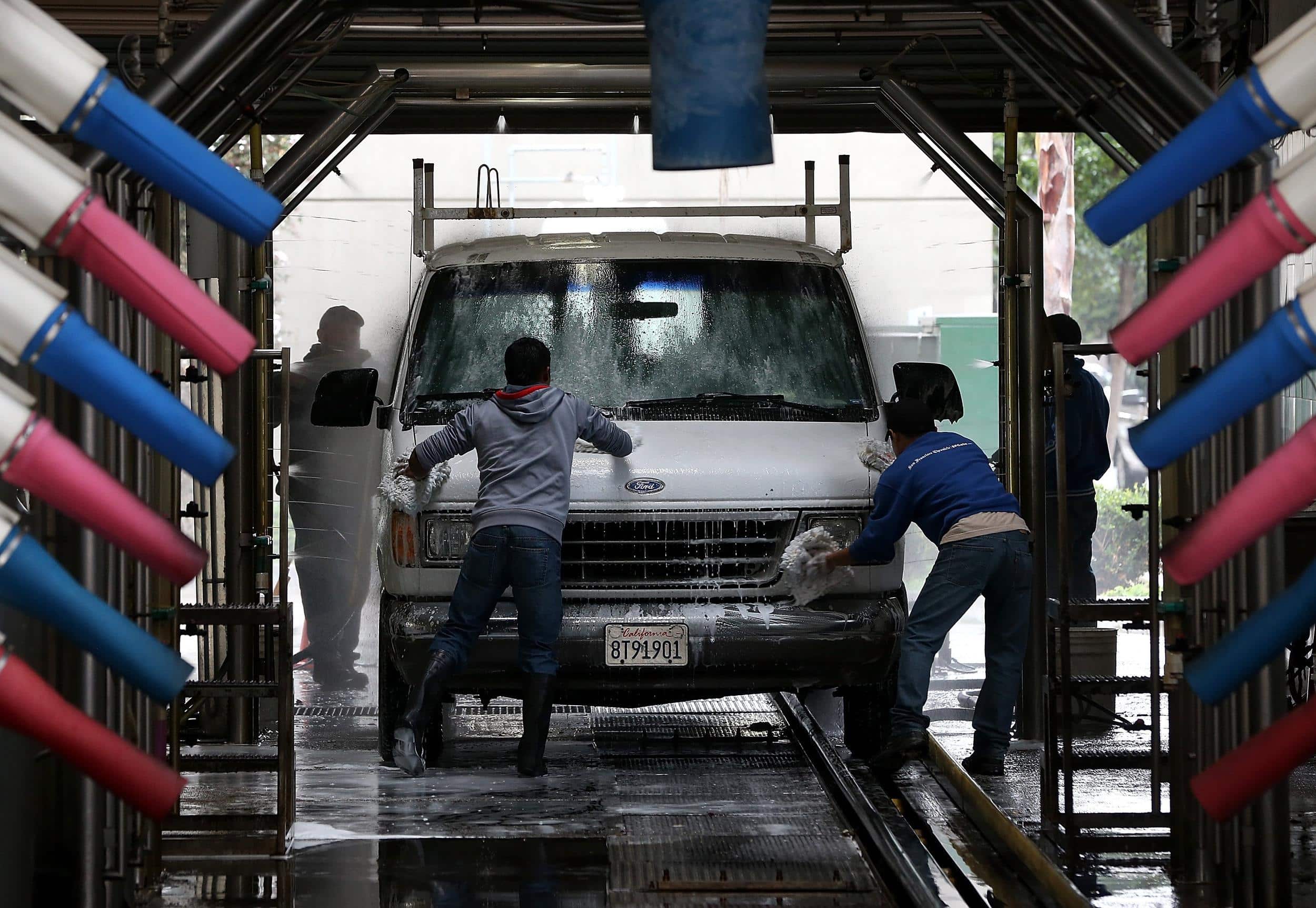 Two individuals are washing a van in an automated car wash with blue and pink foam brushes.