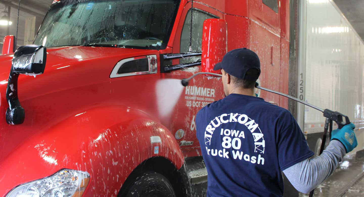 A person is washing a red semi-truck with a high-pressure hose at a truck wash station.
