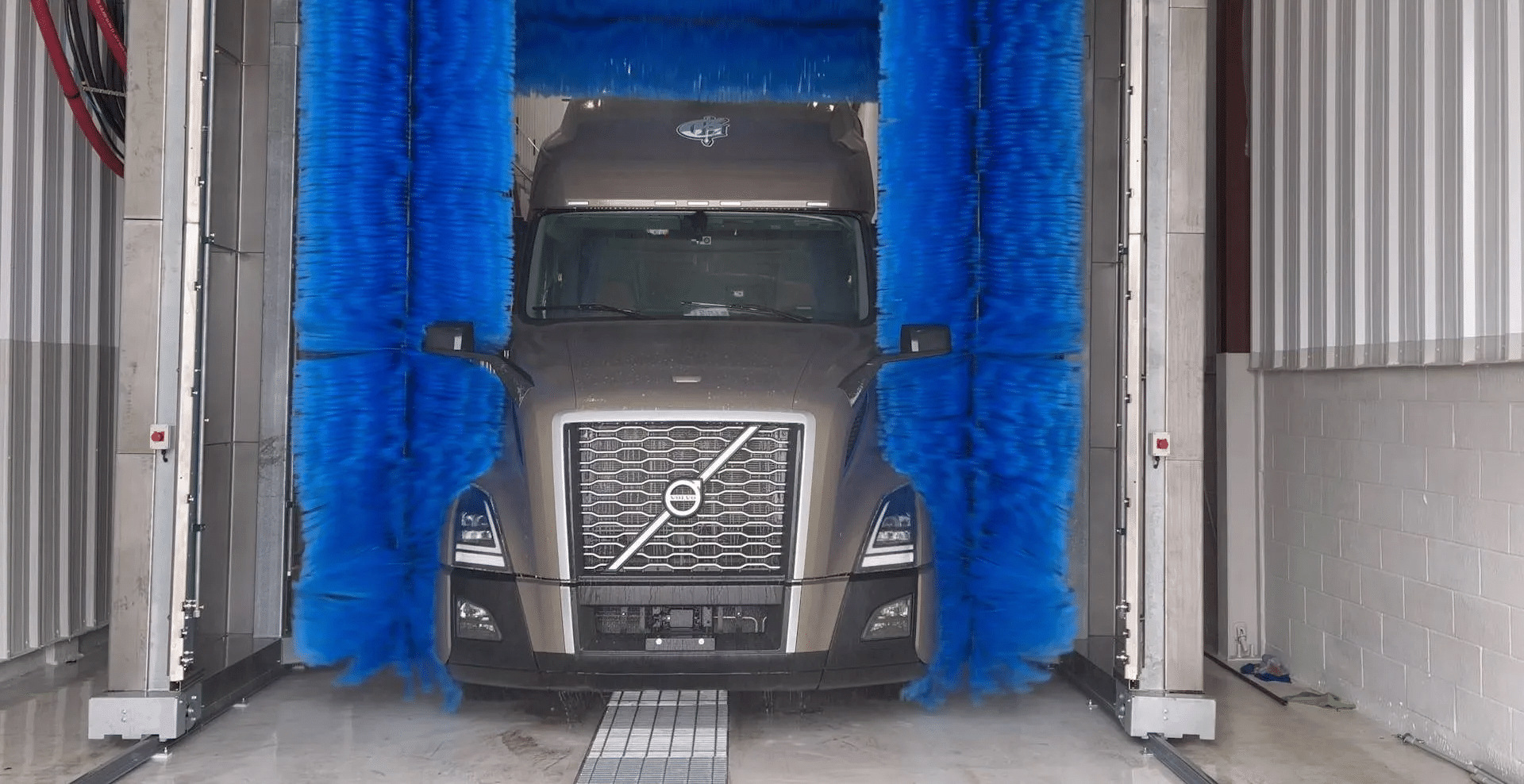 A semi-truck is being washed by large blue brushes in an industrial vehicle wash station.