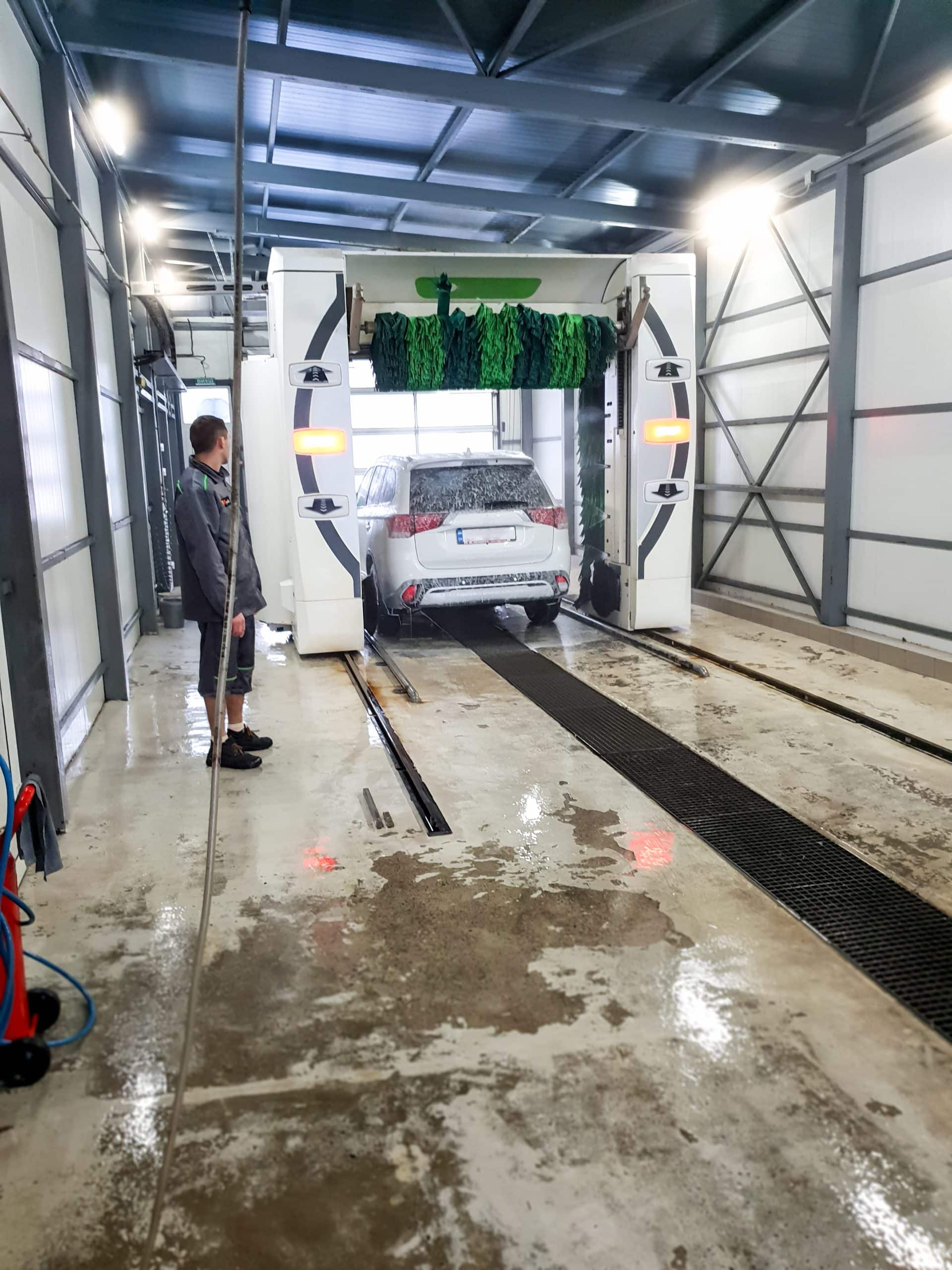 A car is going through an automated wash as an attendant supervises the process inside a wash bay.