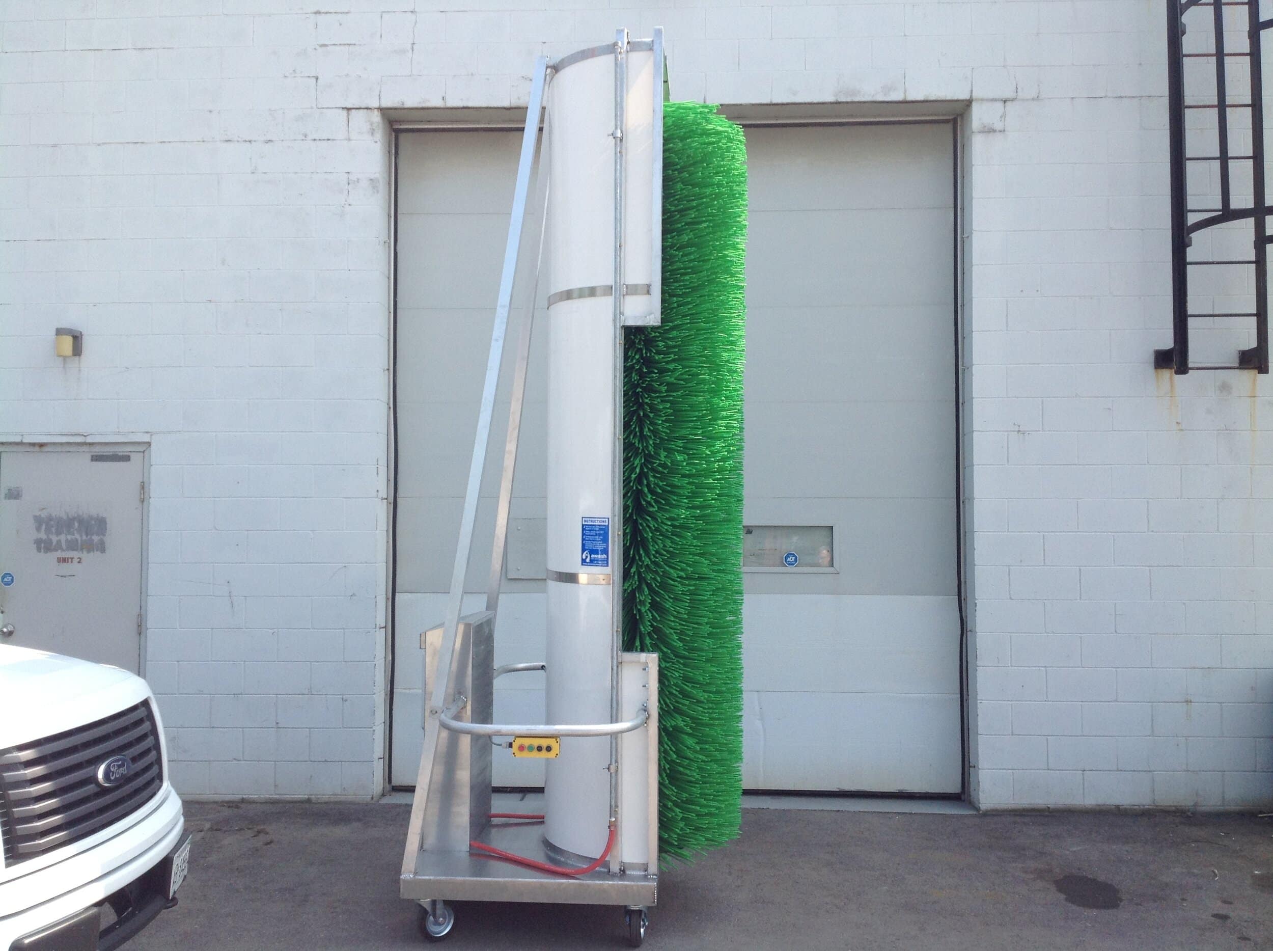 A large cylindrical brush attached to a frame with wheels is stationed in front of a warehouse door.