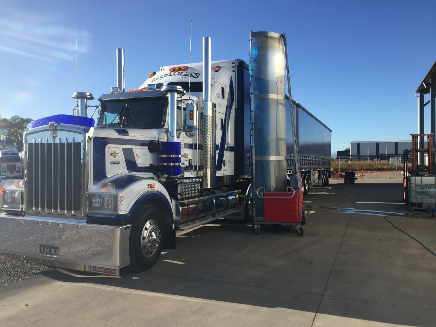 A white semi-truck with blue stripes is parked at a fuel station under a clear sky.