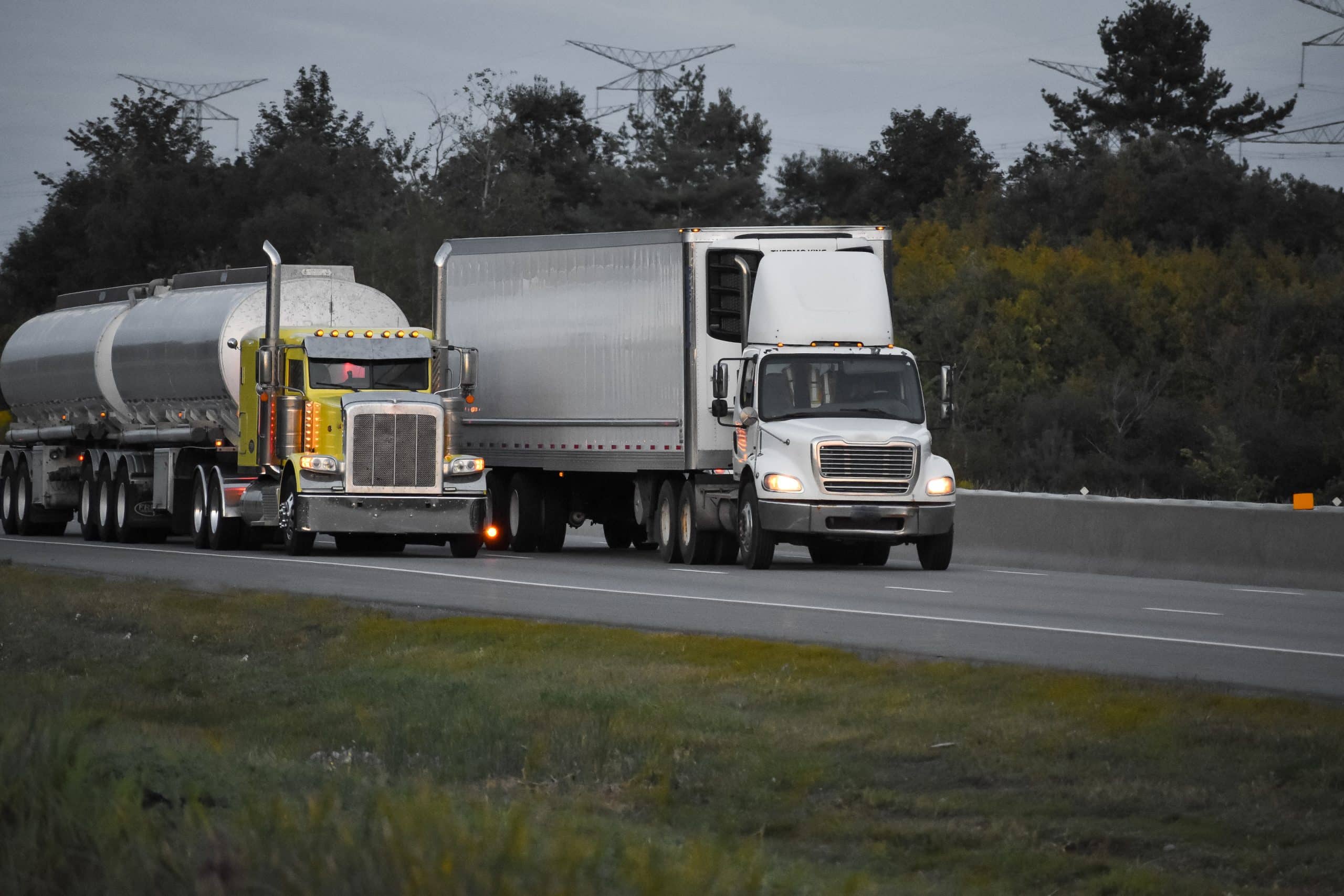 Two large trucks, one with a tanker trailer and another with a dry van, are driving side by side on a highway.