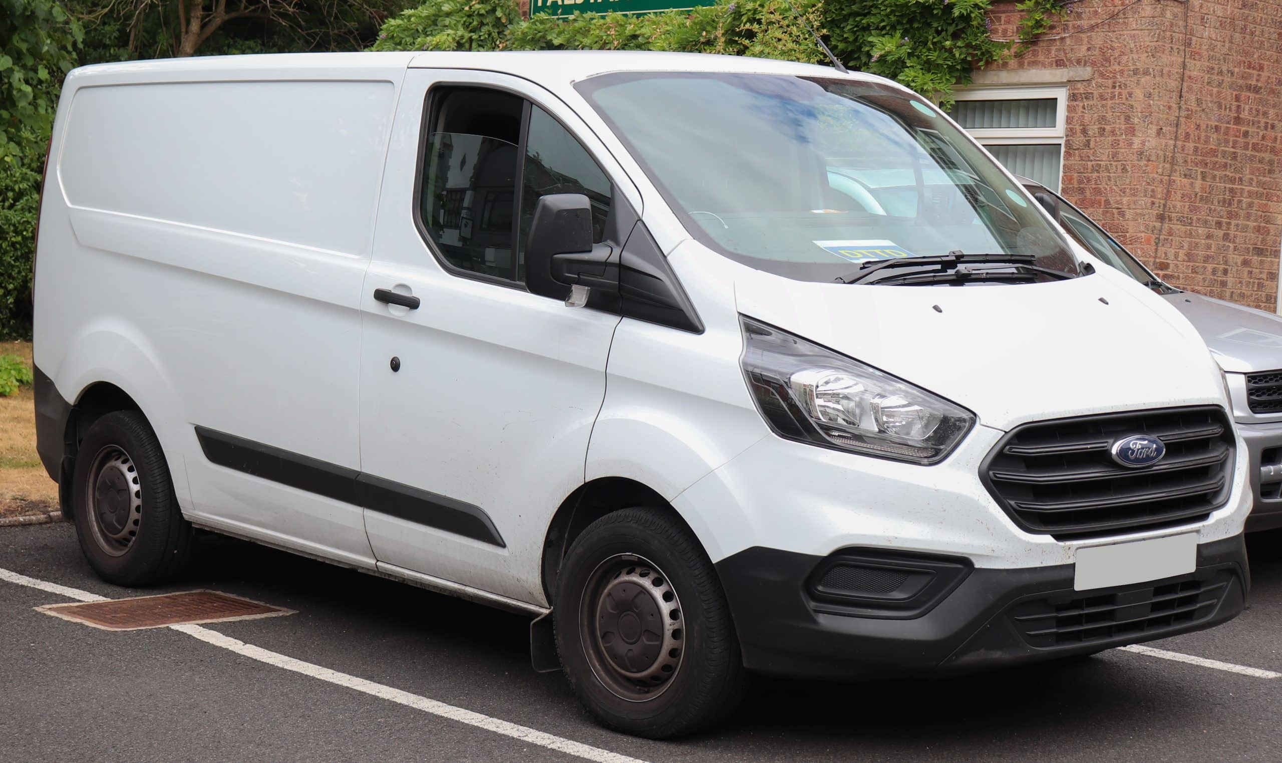 A white Ford commercial van parked at the roadside near a building with foliage in the background.